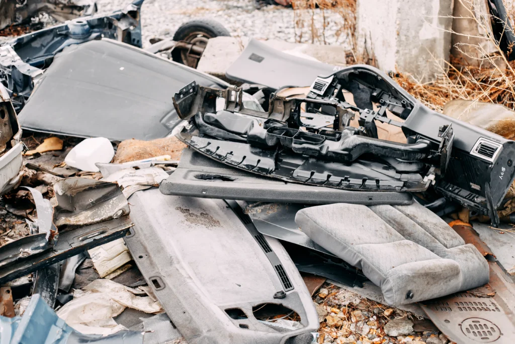 A cluttered pile of discarded car parts, including seats and dashboards, lies on a gravel ground. The scene conveys neglect and abandonment.