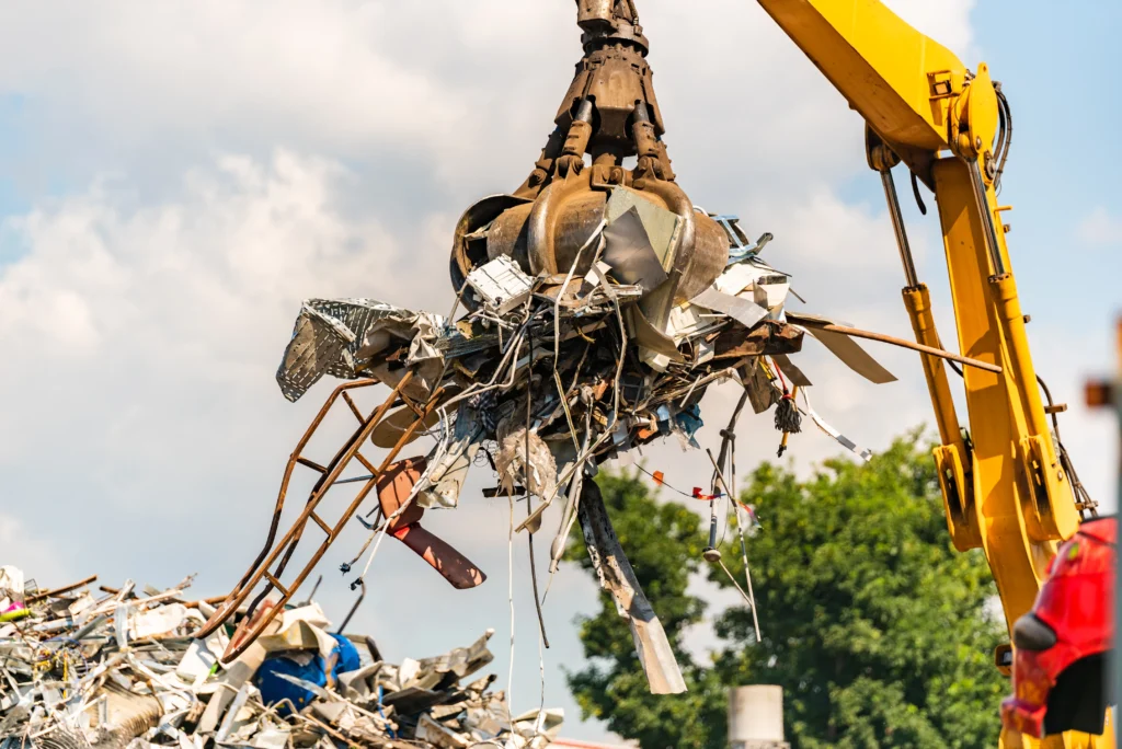 A large crane with a metal claw lifts scrap metal amidst a junkyard. The sky is partly cloudy, and green trees are visible in the background, conveying an industrial yet natural contrast.