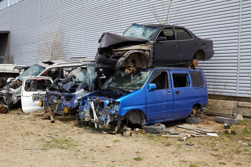 A stack of damaged vehicles in a scrapyard; a black car is atop a blue van with missing parts. The scene conveys abandonment and decay.