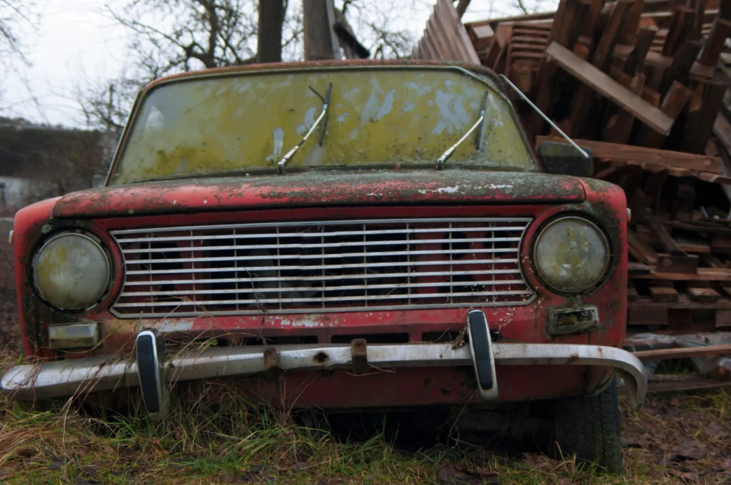 An abandoned, weathered red car with a moss-covered windshield sits in overgrown grass. Its paint is peeling, and wooden pallets are stacked nearby. The scene feels neglected.
